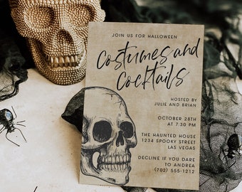 Può includere: Un invito rettangolare di Halloween su carta color cuoio con testo nero Costumes and Cocktails, un'illustrazione di teschio a inchiostro nero, una statuetta di teschio dorata e piccoli ragni di plastica nera.