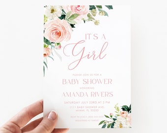 May include: A baby shower invitation with the text "It's a Girl" in pink script. The invitation is decorated with watercolor-style pink and white flowers and green foliage. The invitation is held by a hand.