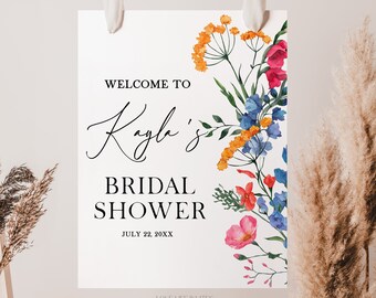 May include: A white rectangular sign featuring the black text "WELCOME TO Kayla's BRIDAL SHOWER JULY 22, 20XX" and a vertical floral arrangement on the right side with orange, pink, and blue watercolor flowers and green leaves.