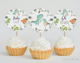 Könnte beinhalten: Drei runde Cupcake-Topper auf Holzstäbchen mit Aquarellillustrationen von grünen und orangefarbenen Dinosauriern und Blättern. Zwei Topper zeigen den Text Baby CRUZ und OH BABY, während der mittlere Topper einen Dinosaurier darstellt.