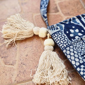 May include: A blue and white patterned tote bag with a matching strap. The bag features cream-coloured tassels and pom-poms. The bag is made of fabric and has a geometric design. The background is a brown, tiled surface.