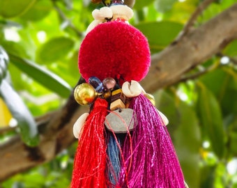 May include: A colorful tassel keychain with a red pom-pom, gold beads, and white shells. The tassel is made of red, pink, and blue yarn.