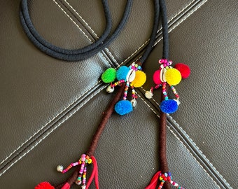 May include: A black cord necklace with colorful pom-poms and beaded accents. The necklace features red, orange, blue, and pink pom-poms, along with small bells and shell details. The cord is dark and the overall design is bohemian.