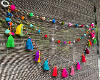 May include: A colourful garland with pom-poms and tassels, strung with beads. The garland is made of various colours, including pink, blue, green, yellow, and red.