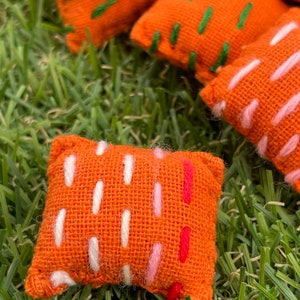 May include: A set of five orange fabric cushions with different coloured embroidery designs. The cushions are on a green grassy surface.