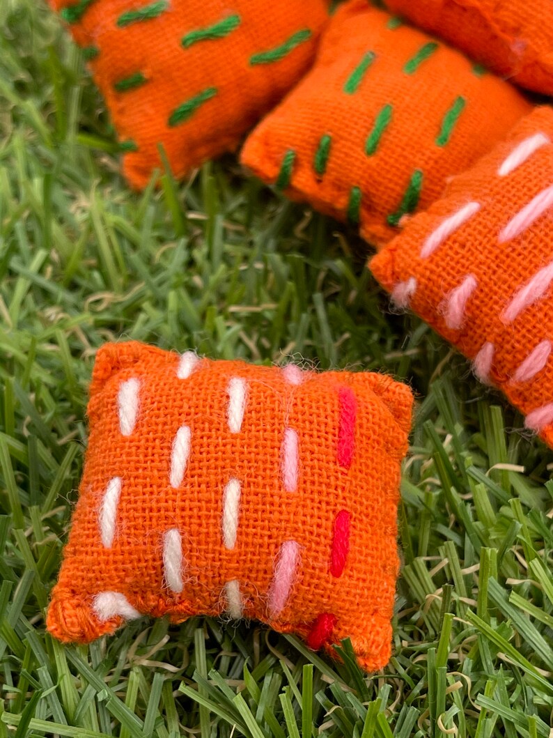May include: A set of five orange fabric cushions with different coloured embroidery designs. The cushions are on a green grassy surface.