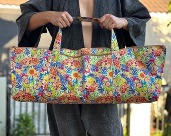 May include: A large, rectangular tote bag with a vibrant floral pattern in shades of yellow, blue, pink, and orange. The bag has brown trim and handles. The bag is held up by a person.