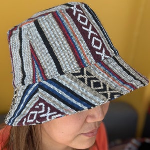 Puede incluir: Un sombrero de pescador con un patrón geométrico colorido en tonos de gris, azul, rojo y blanco. El sombrero está hecho de tejido tejido y tiene un ala ancha.