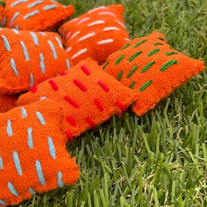 May include: A set of six orange fabric bean bags with colourful embroidered designs. The bean bags are arranged on a green grassy surface.