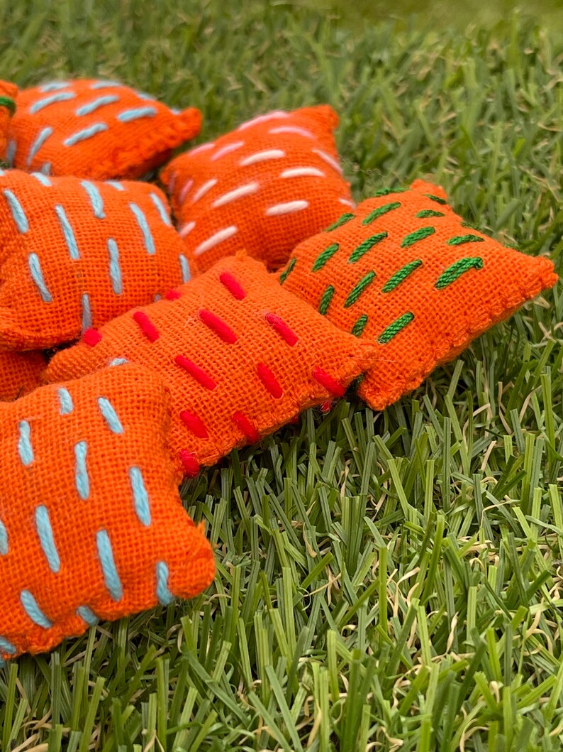 May include: A set of six orange fabric bean bags with colourful embroidered designs. The bean bags are arranged on a green grassy surface.