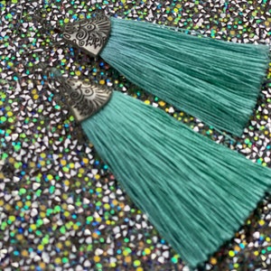 May include: A pair of teal blue tassel earrings with silver filigree accents. The earrings are on a sparkly background.