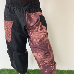 May include: Black cropped pants with a brown and gold patterned design featuring koi fish and floral motifs. The pants have an elastic waistband and a drawstring.