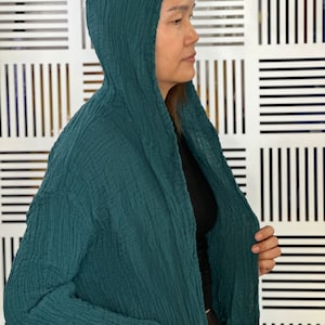 May include: A teal green hooded cardigan with a textured fabric. The cardigan is open in the front and has a long sleeve.
