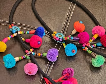 May include: Several black headbands with colorful pom-poms and beaded accents. The pom-poms are in various bright colors, including pink, orange, blue, and purple. The headbands are arranged on a dark surface.