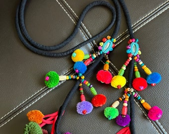 May include: A black cord necklace with colorful pom-poms and beaded accents. The pom-poms are in various colors, including red, purple, green, orange, and yellow. The necklace has a bohemian style.