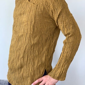 May include: A long-sleeved shirt with a textured, striped pattern in a mustard yellow colour. The shirt has a V-neck and a single pocket on the left side.