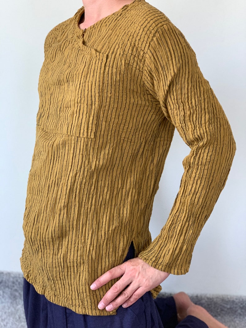 May include: A long-sleeved shirt with a textured, striped pattern in a mustard yellow colour. The shirt has a V-neck and a single pocket on the left side.