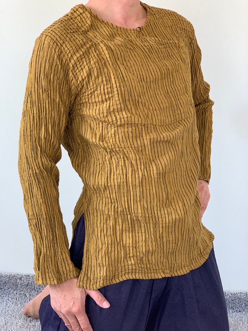 May include: A long-sleeved shirt with vertical stripes in a mustard yellow colour. The shirt has a round neckline and a single button fastening at the neck.