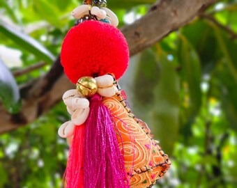 May include: A red and pink tassel keychain with a small fabric pouch decorated with orange and gold embroidery. The keychain is adorned with white seashells and a bell.