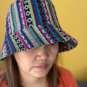 May include: A woman wearing a colorful bucket hat with a striped and geometric pattern. The hat is made of woven fabric and has a wide brim.