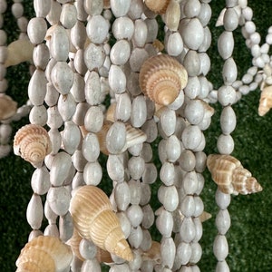 Puede incluir: Una guirnalda de cuentas blancas con conchas colgando de ella. La guirnalda está hecha de pequeñas cuentas blancas y las conchas son de color marrón claro.