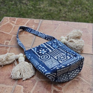 Könnte beinhalten: Eine blau-weiß gemusterte Tragetasche mit passendem Griff und zwei cremefarbenen Quastenakzenten. Die Tasche hat ein geometrisches Design und liegt auf einer braunen Fliesenoberfläche.