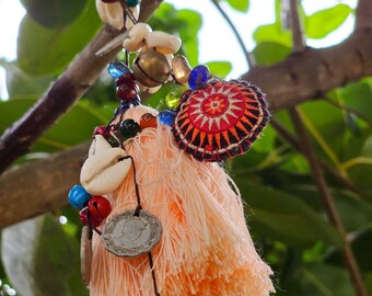 May include: A peach-colored tassel with a bell hanging from it. The tassel is decorated with seashells, beads, and a small embroidered circle with red, orange, and black designs.