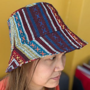 Puede incluir: Un sombrero de cubo colorido con un diseño a rayas y estampado en tonos de rojo, azul, amarillo y blanco.