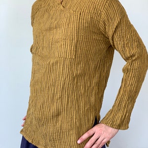 May include: A long-sleeved shirt with a textured, striped pattern in a mustard yellow colour. The shirt has a V-neck and a single pocket on the left side.