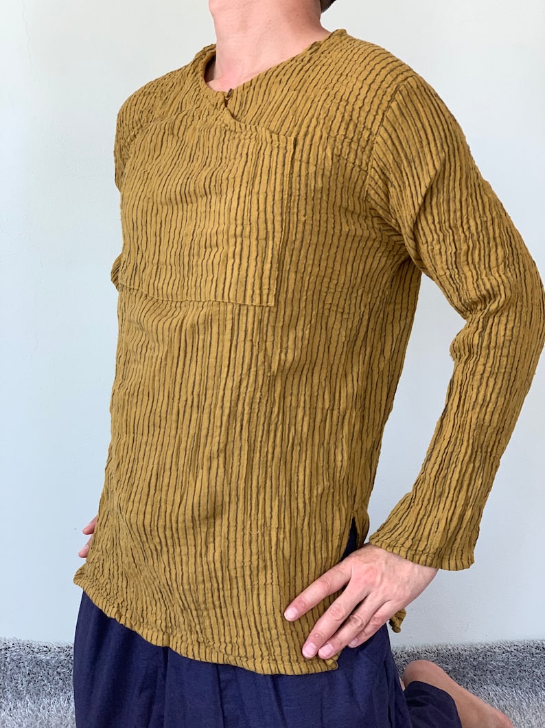 May include: A long-sleeved shirt with a textured, striped pattern in a mustard yellow colour. The shirt has a V-neck and a single pocket on the left side.