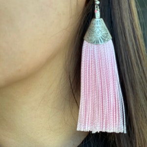 May include: A single pink tassel earring with a silver-colored decorative cap and hook. The earring is made of many strands of pink thread, creating a full, flowing tassel. The earring is shown against a blurred background.