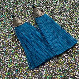 May include: A pair of teal blue tassel earrings with silver metal tops. The tops have a decorative design.