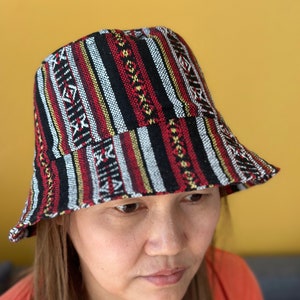 Puede incluir: Un sombrero de cubo a rayas rojas, negras y blancas con un patrón geométrico. El sombrero está hecho de tejido tejido.