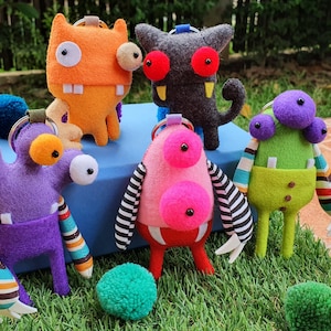 May include: A collection of colorful, handmade monster plush toys. Each plush has unique features, including multiple eyes, teeth, and striped or patterned arms. The monsters are in various colors like orange, purple, green, pink, and gray.