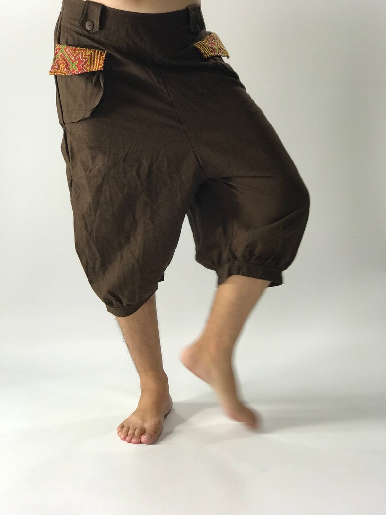 May include: Brown, loose-fitting pants with a decorative embroidered patch on the front pockets. The pants have a drawstring waist and a cuffed hem.