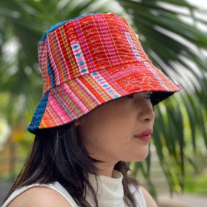 Op de afbeelding: Een kleurrijke bucket hat met een rood, roze, blauw en geel patroon. De hoed is gemaakt van stof en heeft een brede rand.