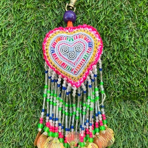 May include: A heart-shaped keychain with intricate embroidery in vibrant colors. The design features a central heart motif with swirling patterns, surrounded by beaded strands in various colors, and adorned with iridescent shell-shaped charms. A gold-tone clasp and purple bead complete the design.