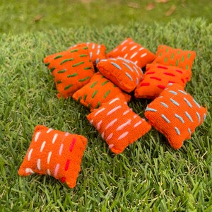 May include: A group of ten small orange throw pillows with different coloured embroidery designs. The pillows are arranged on a green grassy surface.