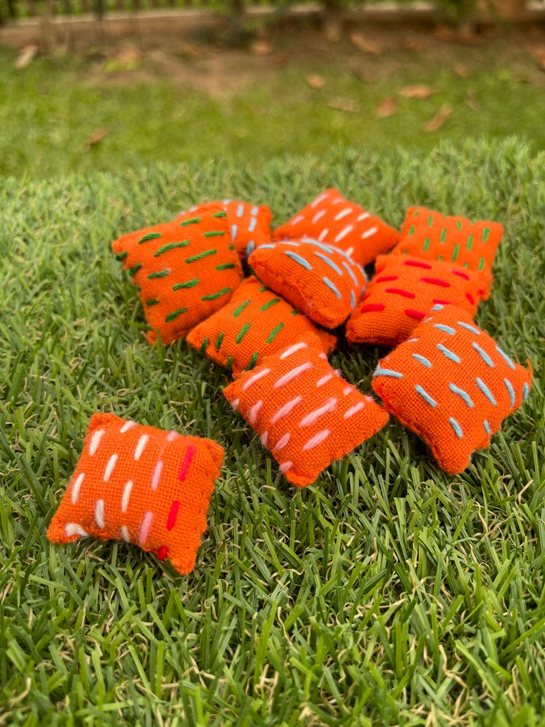 May include: A group of ten small orange throw pillows with different coloured embroidery designs. The pillows are arranged on a green grassy surface.