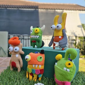 May include: A collection of handmade felt monster toys in various colors. The monsters have large eyes, teeth, and unique features like horns, hats, and striped arms. They are arranged on a green surface with grass and a white, round object in the foreground.