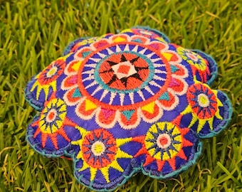 May include: A colourful, round, embroidered cushion with a blue background and intricate patterns in yellow, red, and white. The cushion is sitting on a bed of green grass.