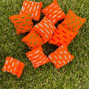 May include: A collection of 10 orange throw pillows with different colored embroidered designs. The pillows are arranged on a green grassy surface.