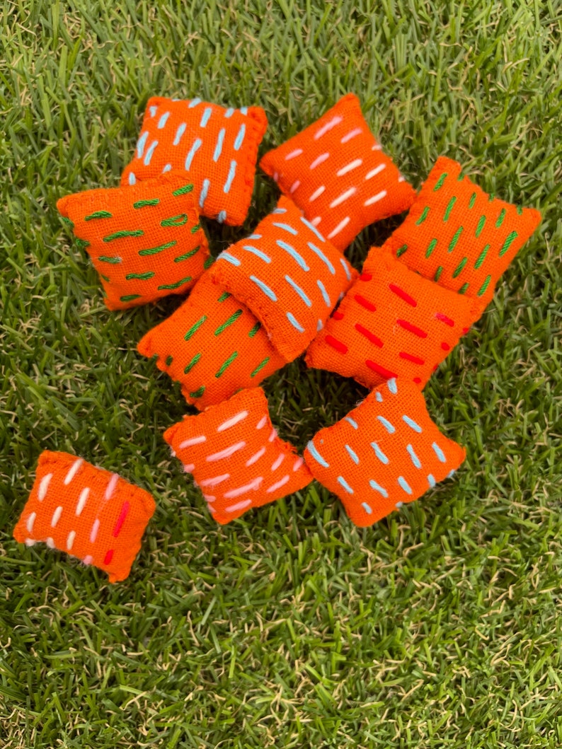 May include: A collection of 10 orange throw pillows with different coloured embroidered designs. The pillows are arranged on a green grassy surface.