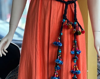 May include: A long, black braided belt with colorful beads and pom-poms hanging from the ends. The belt is tied around a coral-colored dress.