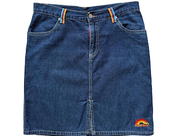 May include: A dark blue denim skirt featuring a front center slit, a metal button closure, and two belt loops accented with vertical rainbow-striped fabric. A small embroidered rainbow logo with the text Billabong is visible at the bottom right.