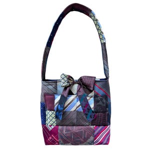 May include: A patchwork handbag made from various patterned neckties. The bag features a rectangular shape with a handle and a decorative bow. The color palette includes shades of blue, purple, gray, and burgundy.