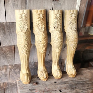 May include: Four antique, carved wooden furniture legs. The legs are a light beige color, with detailed carvings of leaves and grape clusters. They are displayed on a weathered wooden surface.