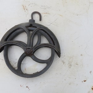 May include: A black cast iron pulley with a decorative design. The pulley has a hook at the top and a wheel with a central hub and spokes.
