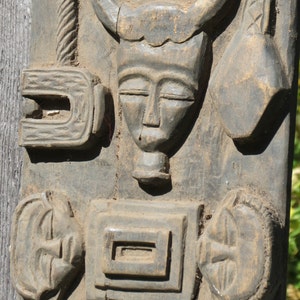 May include: A wooden carving with multiple figures and designs. The carving features a human face with closed eyes, a horned animal head, and a stylized figure with a curved shape. The carving is weathered and has a dark brown color.