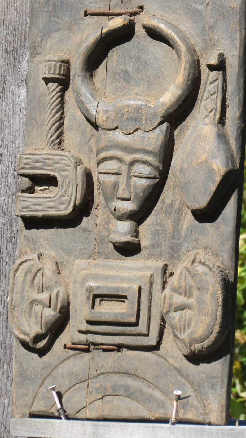 May include: A wooden carving with multiple figures and designs. The carving features a human face with closed eyes, a horned animal head, and a stylized figure with a curved shape. The carving is weathered and has a dark brown color.